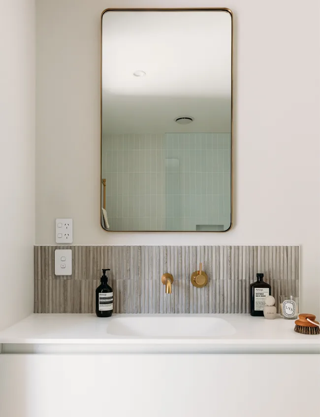 Modern bathroom vanity with a large mirror, gold faucet, and toiletries on a white countertop.