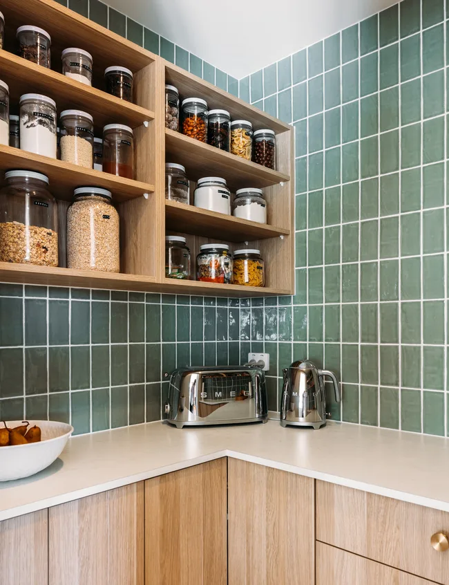 Wooden kitchen shelves with jars, a SMEG toaster, and kettle on a white countertop beside green tiled walls.