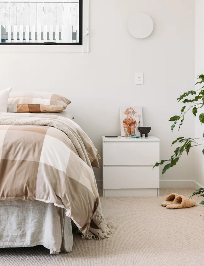 Cozy bedroom with a checkered duvet, white nightstand, artwork, houseplant, slippers, and window with blinds.