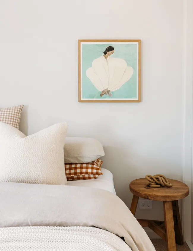 A cozy bedroom with a framed artwork on the wall, neutral bedding, and a wooden side table with a decorative rope.