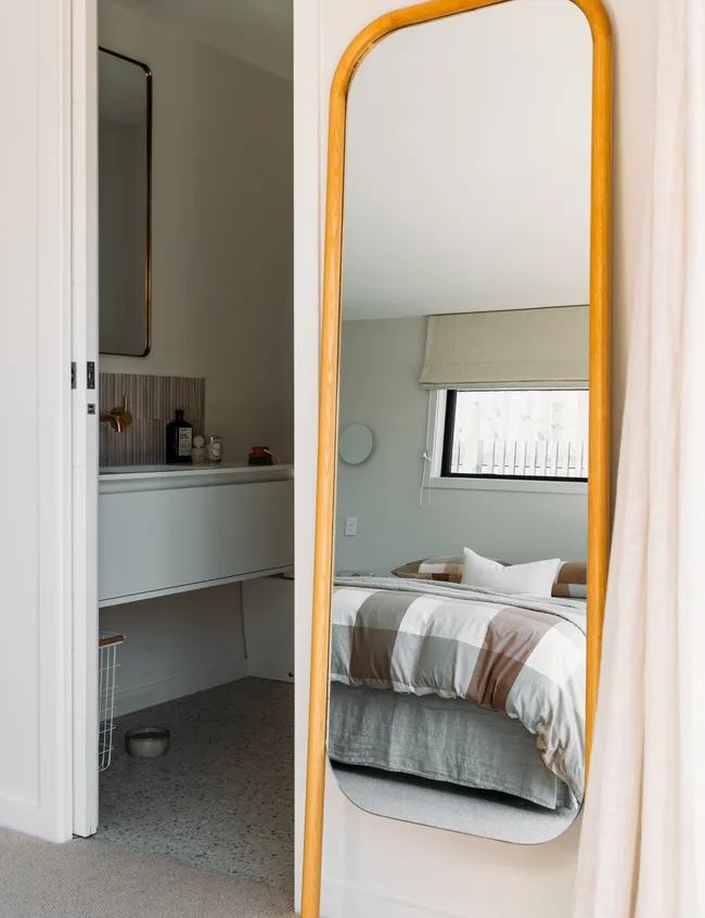 Mirror reflecting a neatly made bed with plaid bedding; bathroom visible with vanity and wall-mounted mirror.