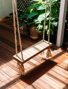 A wooden swing with rope seats hangs on a sunlit wooden deck, surrounded by lush green plants.