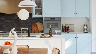 A kitchen painted light blue with a white pendant light, champagne on the bench and a coffee machine.