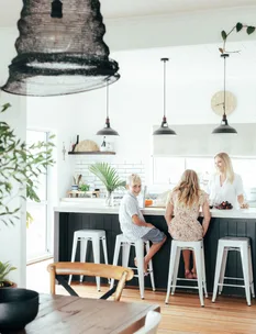 Three people sit at a kitchen island with hanging lights and indoor plants in a modern, bright kitchen.