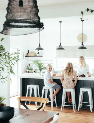 Three people sit at a kitchen island with hanging lights and indoor plants in a modern, bright kitchen.