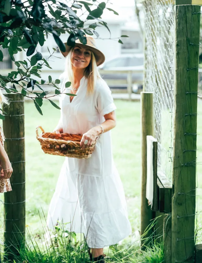 Woman in white dress and hat holds a basket, smiling outdoors near a fence with greenery.