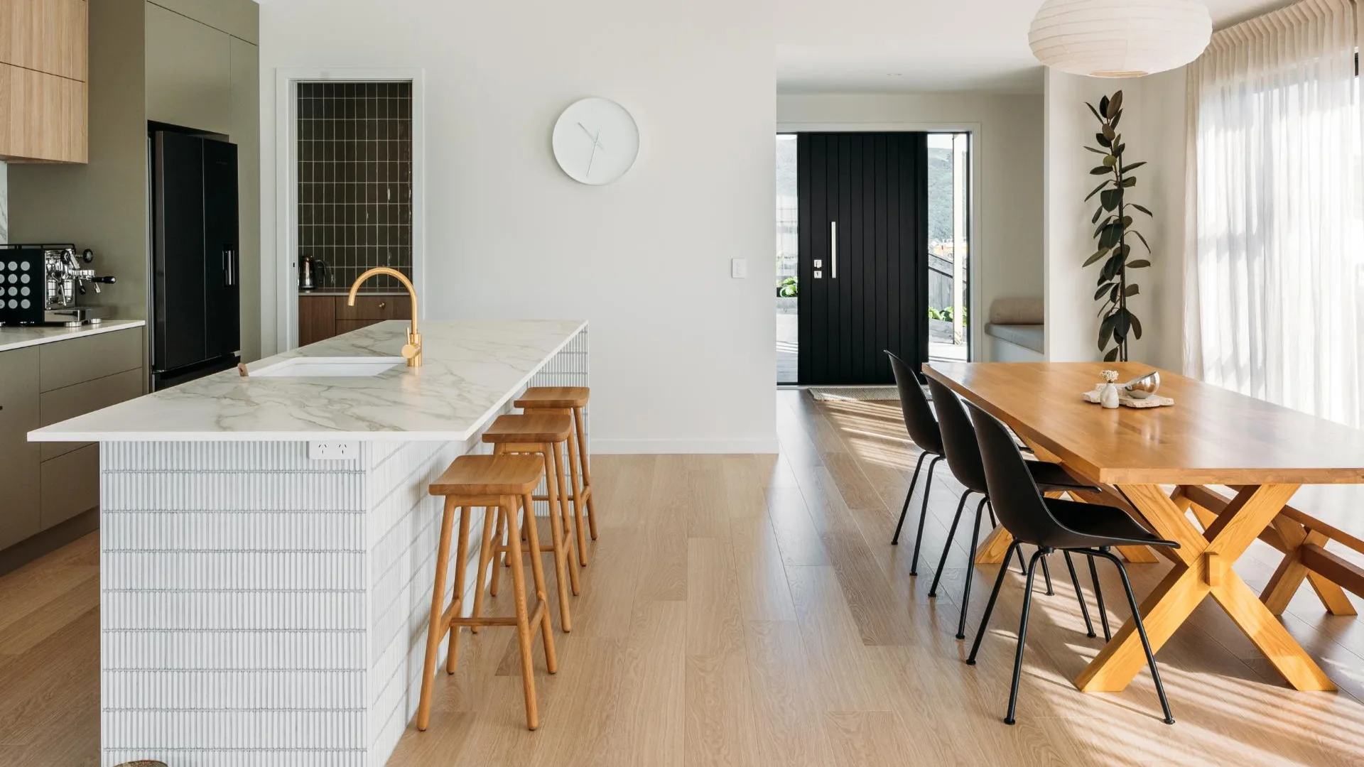 Modern kitchen and dining area with a marble island, wooden stools, a wooden table, black chairs, and natural light.