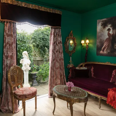 Opulent room with green walls, ornate furniture, a garden view, and classical decor including a bust and painting.