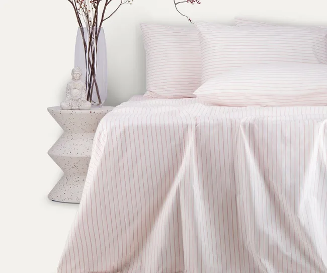 Bed with white and pink striped sheets, pillows, a side table with a vase, branches, and a small Buddha statue.