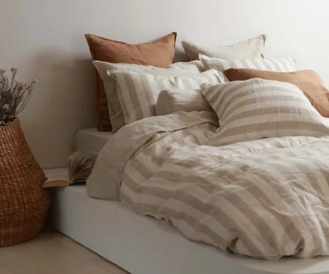Cozy bed with striped linens, several pillows, and an open book beside a wicker basket with dried flowers.