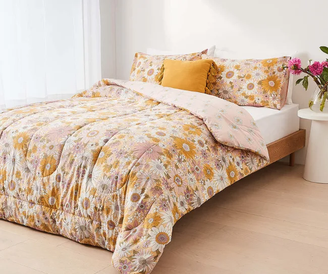 Floral-patterned comforter set on a wooden bed with orange accent pillow and pink flowers on a bedside table.
