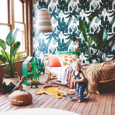 Toddler in a lively room with tropical wallpaper, toy dinosaurs, a skateboard, fur rug, and plant decor.