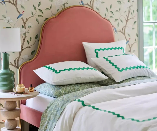 Pink upholstered bed with green and white bedding, next to a lamp and small table, floral wallpaper in the background.