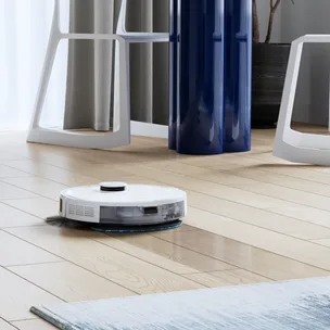 A white robotic vacuum cleaner on a wooden floor near modern furniture and a dark potted plant.