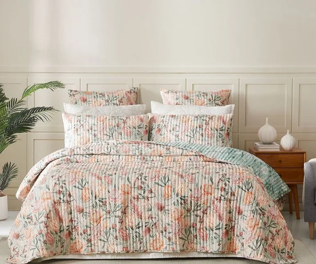 Floral quilted bedding set with pillows and blanket on a bed, with a plant and nightstand in the background.