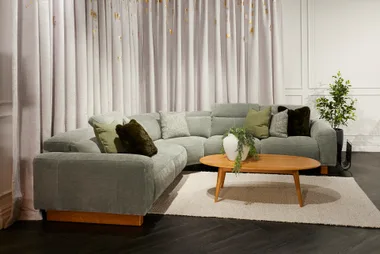 Cozy living room with a grey sectional sofa, green pillows, wooden coffee table, and light curtains.