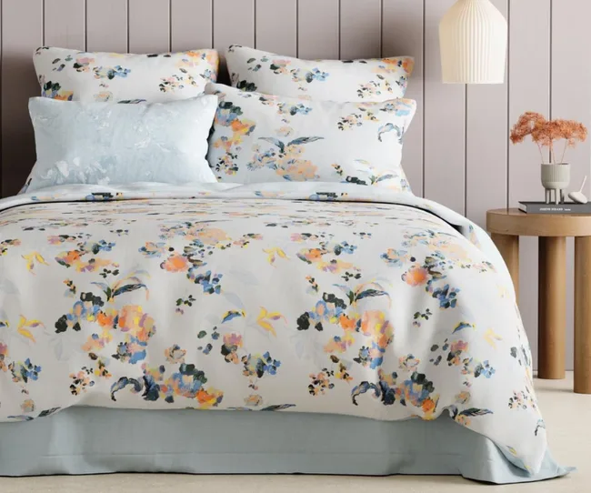 Floral-patterned bedding with pillows on a bed in a room with muted pink walls and a side table with decor items.