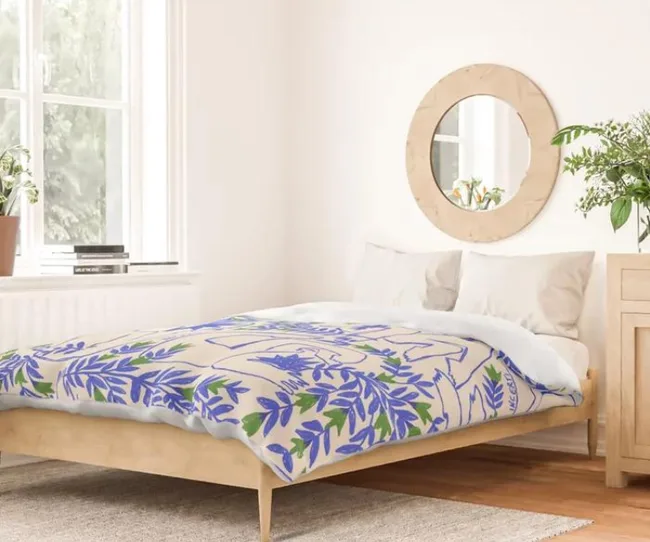 Cozy bedroom with a wooden bed, floral bedding in blue and green, round mirror, and potted plants by the window.