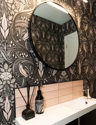 Bathroom with ornate wallpaper, round mirror, soap dispenser, and diffusers on a white sink.