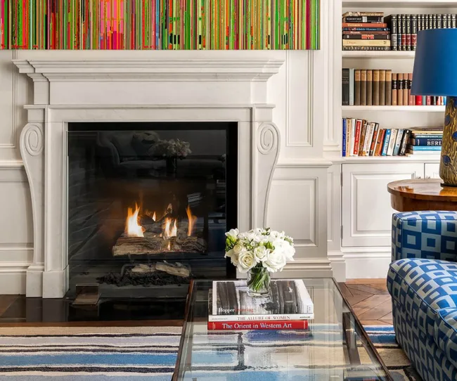 Fireplace with colorful art above, bookshelves on the side, and a glass table with flowers and books in the foreground.
