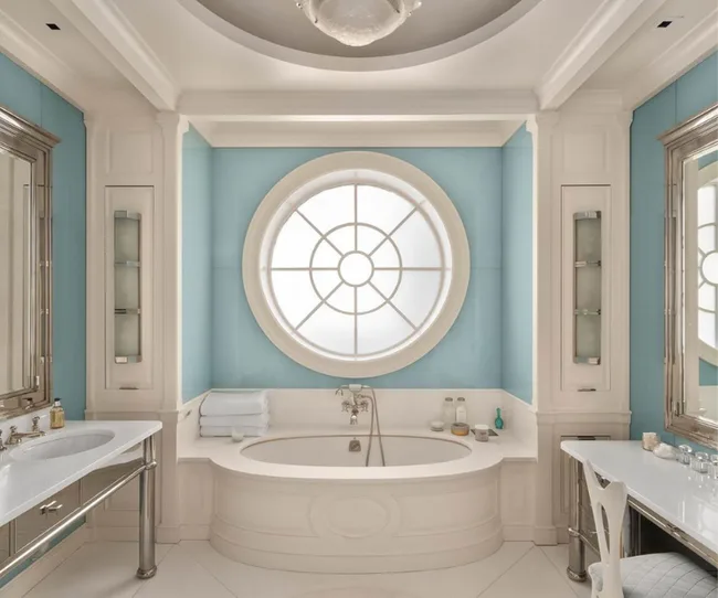 Elegant bathroom with a circular window, white tub, blue walls, and silver-framed mirrors.