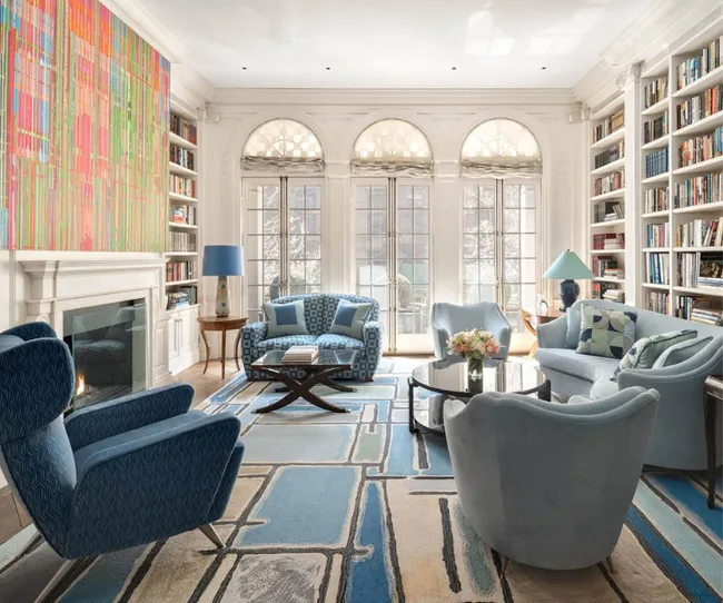Luxurious living room with blue armchairs, colorful artwork, large bookshelf, and arched windows.
