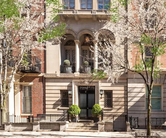 Historic townhouse facade with arched windows, ornate details, blooming trees, and a sunny sidewalk.