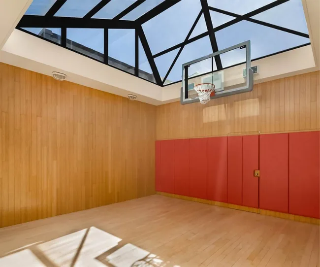 Indoor basketball court with wooden floors, red padded walls, and a glass ceiling with natural light.