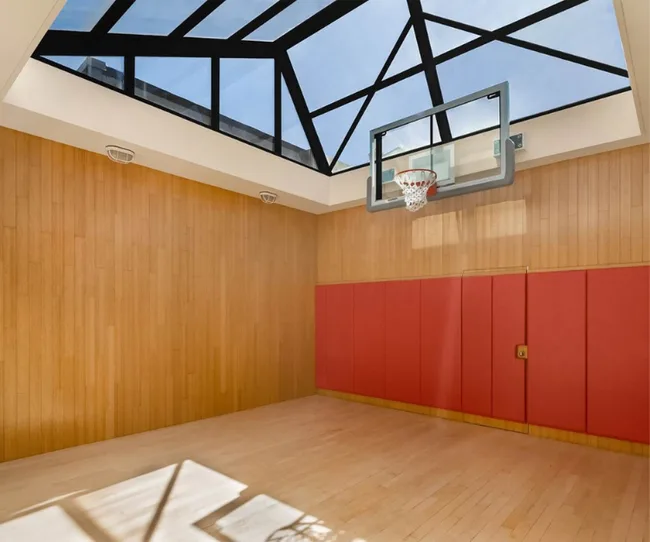 Indoor basketball court with wooden floors, red padded walls, and a glass ceiling with natural light.