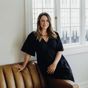 Woman in a black dress leaning on a brown couch in front of a window with geometric patterns.