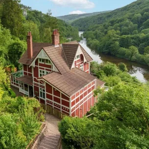 Red and white Tudor-style house by a river, surrounded by lush green trees and hills.