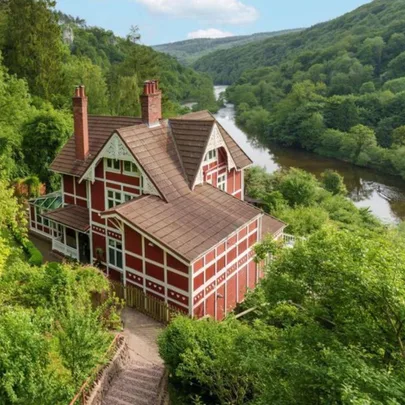 Red and white Tudor-style house by a river, surrounded by lush green trees and hills.
