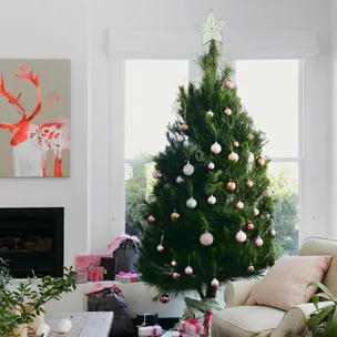 Christmas tree with pink and gold ornaments, star topper, gifts beneath, next to a reindeer painting and armchair.