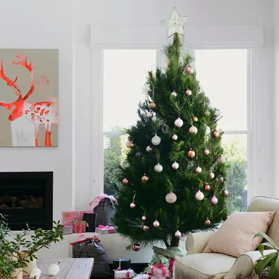 Christmas tree with pink and gold ornaments, star topper, gifts beneath, next to a reindeer painting and armchair.
