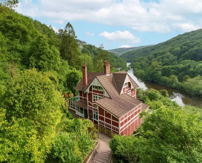 Red and white house with a sloped roof, surrounded by lush green trees, overlooking a river and hills under a partly cloudy sky.