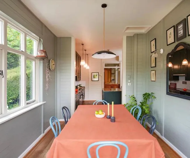 Dining room with orange tablecloth, blue chairs, green walls, window view, plants, and pendant lights.