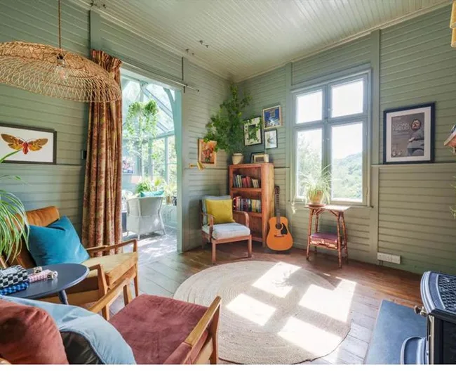 Cozy sunlit room with green walls, guitar, bookshelf, and plants; vintage decor with wicker lamp and round rug.
