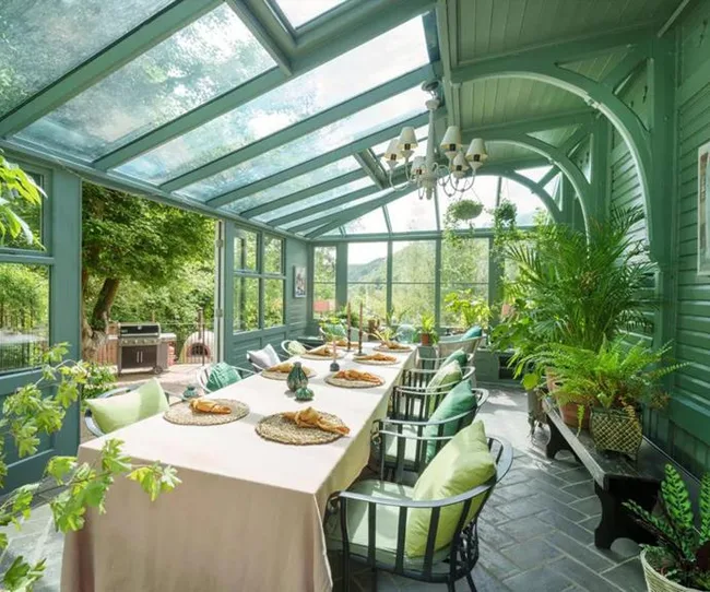 A bright green conservatory with a long table, chairs, and lush plants under a glass roof.