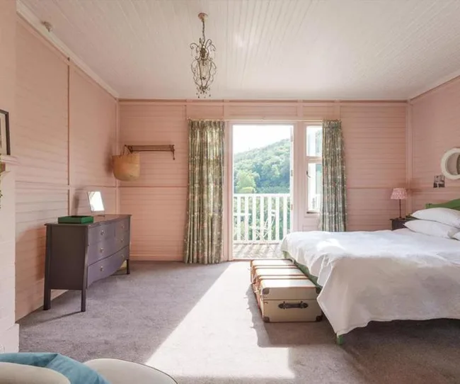 A cozy bedroom with pink walls, a bed, and a dresser. The window overlooks a green landscape with curtains slightly open.
