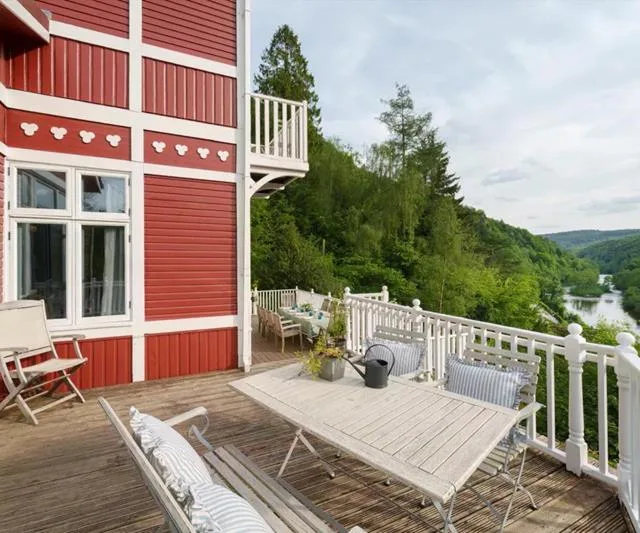 Red and white house with wooden deck, chairs, and tables overlooking lush green trees and a river.