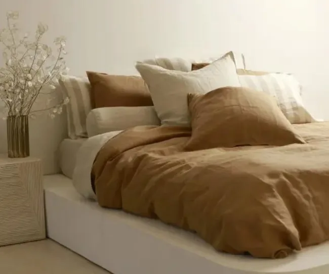 A bed with brown and cream pillows and comforter, next to a wooden nightstand with a vase of dried flowers.