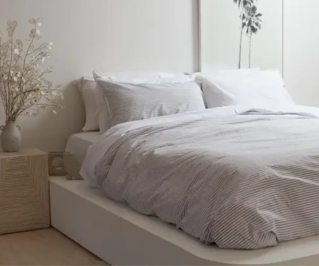 Minimalist bedroom with a striped duvet, white pillows, wooden nightstand, and a vase holding delicate flowers.
