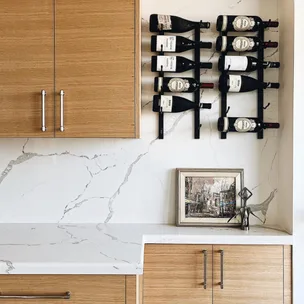 Wine bottles on a wall rack above a marble countertop with light wood cabinets and a framed picture.