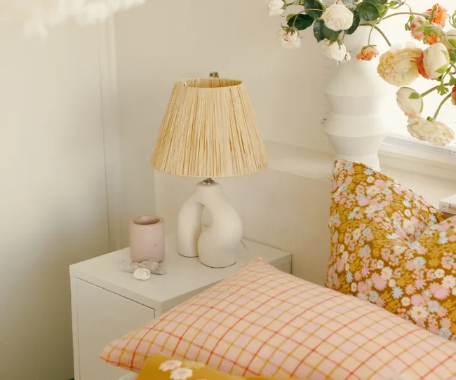 Bedroom scene with a floral pillow, a bedside table holding a modern lamp, a pink cup, and a vase of flowers.