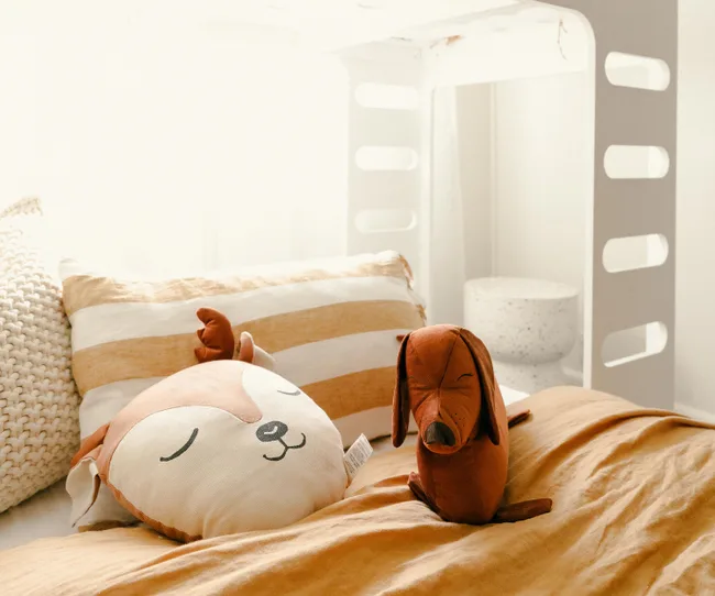 Plush dog and fox face cushions on a cozy bed with striped pillow and warm-toned bedding in a bright room.