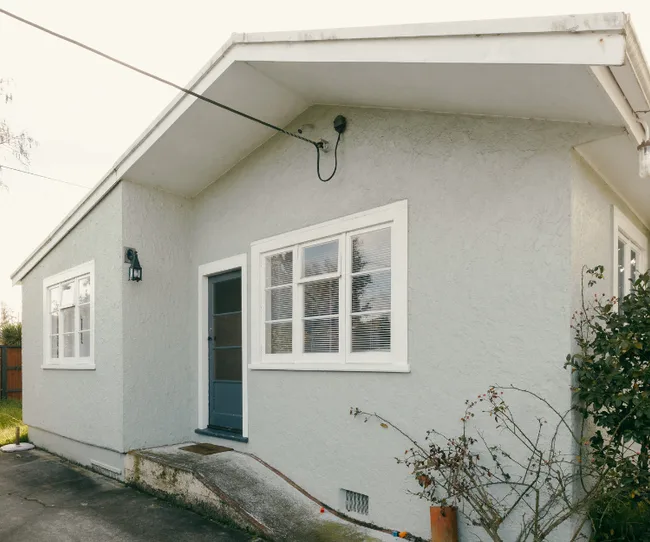 Exterior of a small, light gray house with a blue door, white framed windows, and a concrete path.