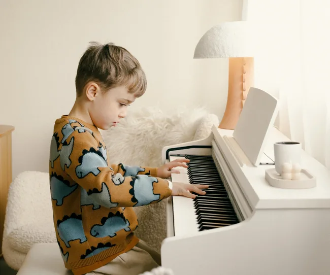 Child wearing a dinosaur sweater plays a white piano in a cozy room.