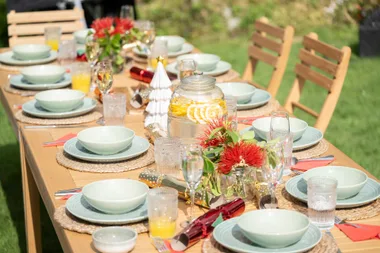Outdoor dining table set with plates, glasses, a jar of lemon water, red flowers, and festive decorations.