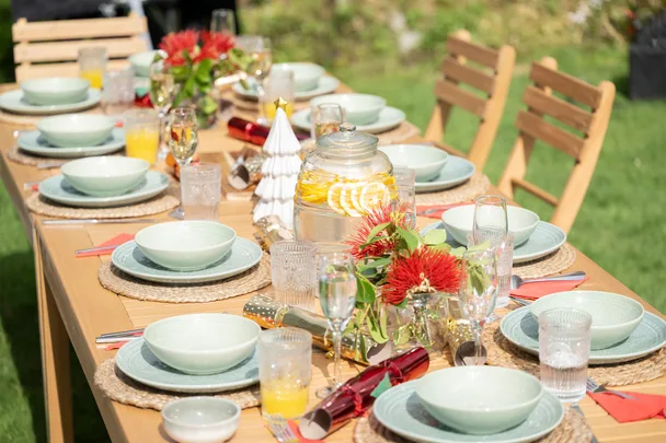Outdoor dining table set with plates, glasses, a jar of lemon water, red flowers, and festive decorations.