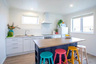 Modern kitchen with white cabinets, colorful stools, fruit bowl, and plants on a wooden island table.